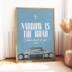 以下が含まれることがあります： 淡い青色の背景に「NARROW IS THE ROAD That leads to life」というテキストと十字架が描かれた額入りのプリント。下部にはヴィンテージの青い車があります。フレームは明るい茶色です。