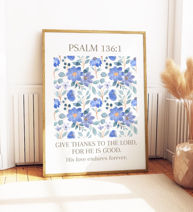 Op de afbeelding: Ingelijste kunstdruk met een bloemenpatroon van blauwe bloemen en groene bladeren op een witte achtergrond. De tekst "PSALM 136:1" staat bovenaan, met het citaat "GIVE THANKS TO THE LORD, FOR HE IS GOOD. His love endures forever." eronder.