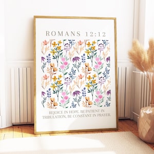 May include: A framed print featuring a floral pattern in watercolor style. The design includes pink, yellow, purple, and blue flowers and foliage. The text "ROMANS 12:12" is at the top, with a quote below: "REJOICE IN HOPE. BE PATIENT IN TRIBULATION, BE CONSTANT IN PRAYER."