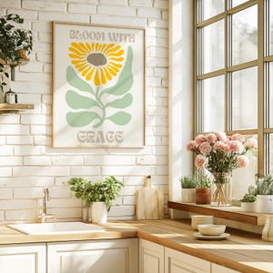 May include: A framed art print with a sunflower design and the words "BLOOM WITH GRACE" in a kitchen setting. The sunflower has yellow petals and a brown center, with green leaves and a light background.