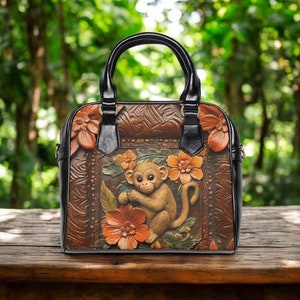 May include: Brown leather handbag featuring a monkey relief design with orange flowers. The bag has a black handle and trim. The background is a blurred green, suggesting a natural setting.