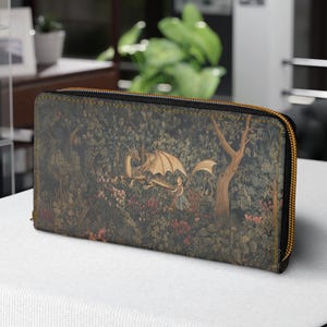 Pode incluir: Uma carteira retangular com zíper dourado e uma cena detalhada de floresta com dragões. O design da carteira inclui uma variedade de cores, incluindo verde, marrom e dourado. A carteira é colocada em uma superfície branca.