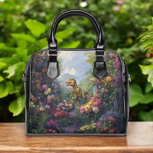 May include: Black handbag with a colorful floral and dinosaur print. The bag has a black handle and trim. The artwork depicts a dinosaur in a garden with colorful flowers and a castle in the background.