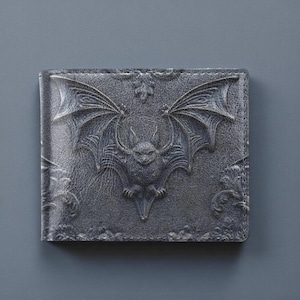 Peut inclure: Un portefeuille gris plié en deux avec un motif de chauve-souris en relief sur le devant. La chauve-souris a des ailes déployées et est centrée sur le portefeuille. Le portefeuille a un aspect texturé et une bordure décorative.