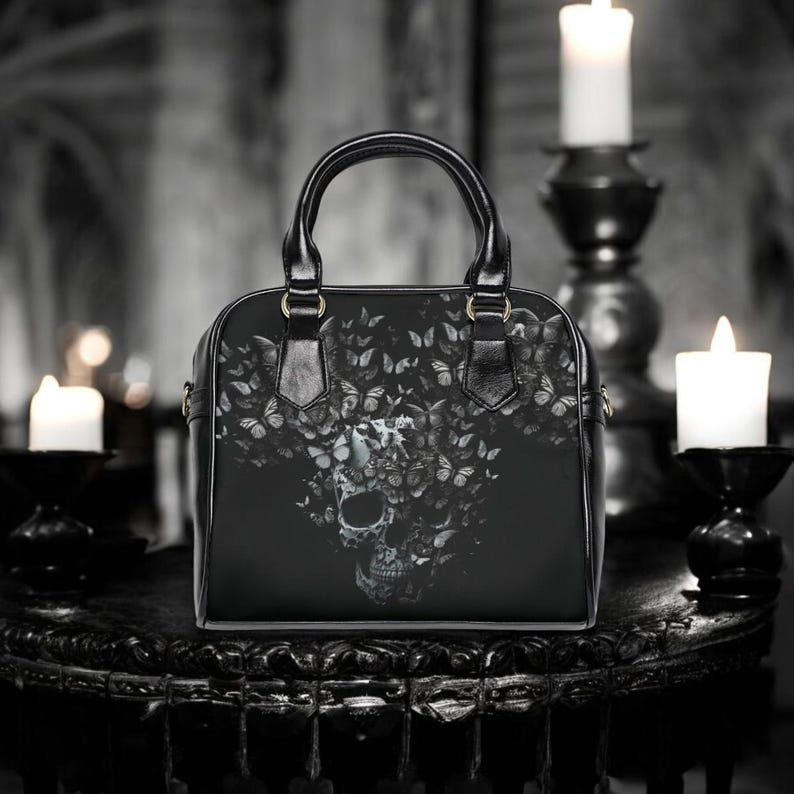 May include: Black handbag featuring a skull design composed of butterflies. The bag has a structured shape with a top handle and a shoulder strap. The skull and butterfly graphic is in shades of gray and white against a black background.