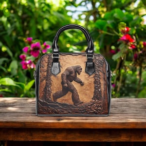 May include: A brown leather handbag with a black handle and trim. The bag features a detailed embossed image of Bigfoot walking through a forest scene. The bag is sitting on a wooden surface.