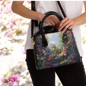 May include: Black satchel bag with a colorful floral and dinosaur print. The bag has a black top handle and a long, adjustable shoulder strap. The image on the bag features a dinosaur in a garden setting with colorful flowers.
