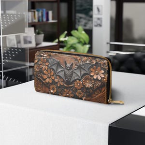 May include: A brown leather wallet with a bat and floral design. The wallet features a detailed embossed pattern with a central bat figure and surrounding flowers. It has a gold-colored zipper.
