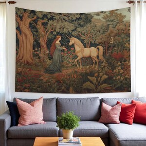 May include: A tapestry depicting a woman in a blue dress and a unicorn in a forest setting. The artwork features a detailed landscape with trees, flowers, and a soft color palette of greens, browns, and creams. The tapestry is hung above a gray sofa with red and pink pillows.