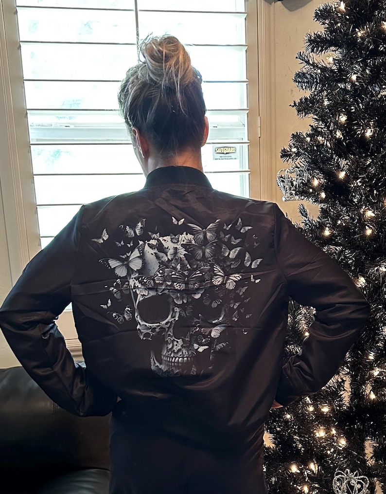 May include: Black bomber jacket featuring a skull design composed of butterflies. The skull is in shades of gray and white, with the butterflies in various sizes and patterns. The jacket has a classic bomber style with a ribbed collar and cuffs.