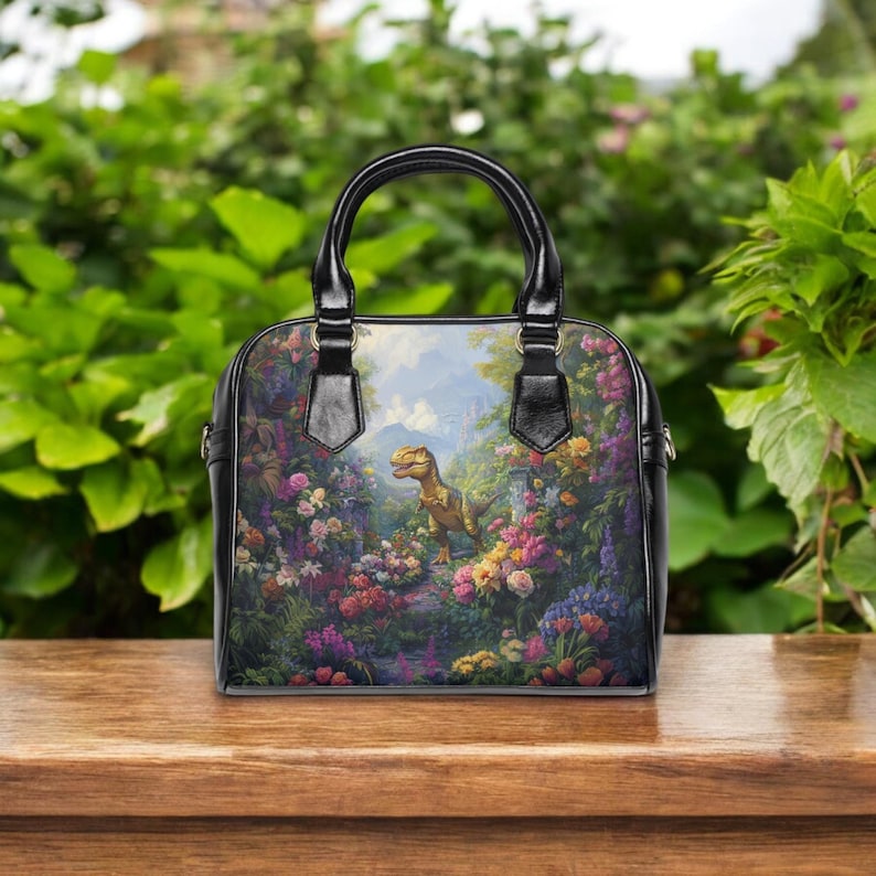 May include: Black handbag with a colorful floral and dinosaur print. The bag has a black handle and trim. The artwork depicts a dinosaur in a garden with colorful flowers and a castle in the background.
