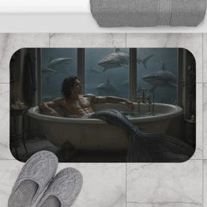 May include: A rectangular bath mat featuring a print of a person with a mermaid tail sitting in a clawfoot bathtub. The scene shows a large window looking out into deep water filled with several sharks. The mat has a gray, textured surface.