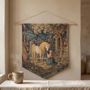 Könnte beinhalten: Ein rechteckiger Wandteppich mit einem Einhorn und einer Frau in einer Waldszene. Der Wandteppich wird an einer Holzstange mit einer Schnur aufgehängt. Die Farben sind gedämpft, mit Grün-, Blau- und Brauntönen, die das Bild dominieren.