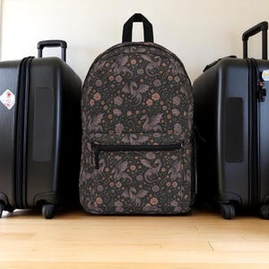 May include: A black backpack with a dragon and floral pattern, featuring a front zipper pocket and a top handle. The backpack is flanked by two black rolling suitcases. The design includes dragons, roses, and other flowers.