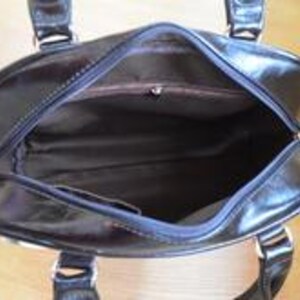 May include: A black, open handbag with a glossy finish. The interior is visible, revealing a brown lining and a small pocket. The bag has two handles and silver-colored hardware. The bag is sitting on a light-colored wooden surface.