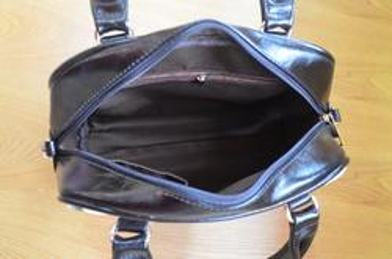 May include: A black, open handbag with a glossy finish. The interior is visible, revealing a brown lining and a small pocket. The bag has two handles and silver-colored hardware. The bag is sitting on a light-colored wooden surface.