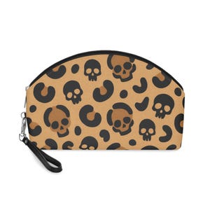 May include: A tan cosmetic bag with a black zip and wrist strap. The bag has a repeating pattern of black skulls and leopard spots. A unique accessory for carrying makeup or other small items.
