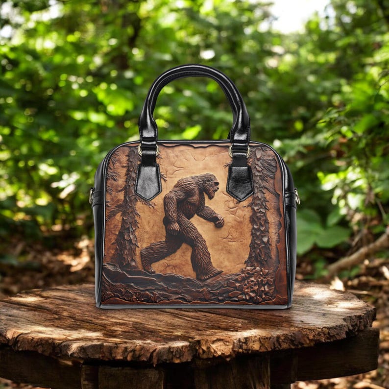 May include: A brown handbag with a black handle and trim. The bag features a detailed relief of a Bigfoot figure walking through a forest scene. The bag is sitting on a wooden surface.