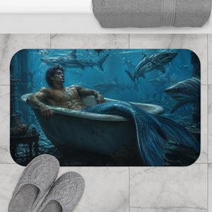 Puede incluir: Una alfombrilla de baño rectangular con una ilustración digital de un tritón con una larga cola azul sentado en una bañera blanca con patas sumergida bajo el agua. La escena incluye varios tiburones nadando en las profundidades del océano.