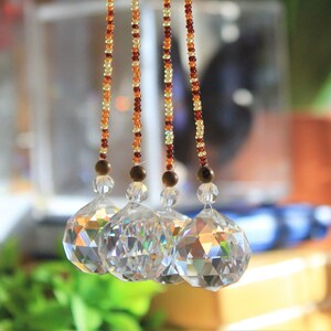 May include: Close-up of a decorative hanging crystal prism. The prism is faceted and clear, reflecting light. Strands of small, colorful beads in shades of gold, amber, and brown lead to the prism. The background is blurred, with hints of green.