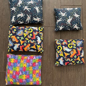 May include: Several decorative cushions with diverse patterns. Designs feature astronauts on a navy background, colourful dinosaurs and rainbows, and a jigsaw puzzle pattern. The cushions are square and rectangular, arranged on a wooden floor.