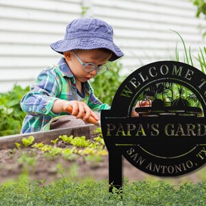 K&ouml;nnte beinhalten: Schwarzes Garten Schild aus Metall mit dem Text "Willkommen im Garten von Papa - San Antonio, TX". Das Schild zeigt eine Silhouette eines Gem&uuml;sekorbs. Ein kleines Kind arbeitet im Hintergrund im Garten.