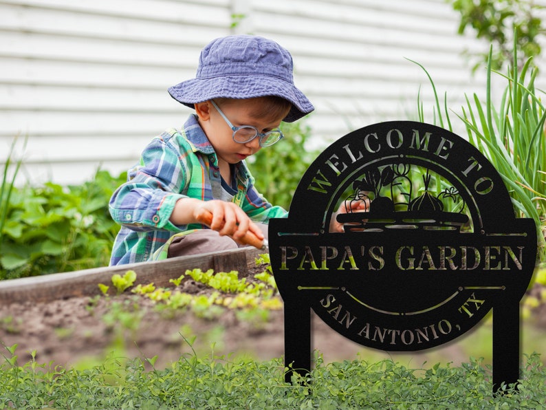 K&ouml;nnte beinhalten: Schwarzes Garten Schild aus Metall mit dem Text "Willkommen im Garten von Papa - San Antonio, TX". Das Schild zeigt eine Silhouette eines Gem&uuml;sekorbs. Ein kleines Kind arbeitet im Hintergrund im Garten.