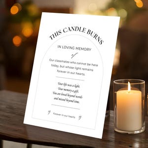 May include: A white card with the text "THIS CANDLE BURNS IN LOVING MEMORY" and a poem, displayed next to a lit pillar candle in a glass holder. A second candle in a jar is also visible.