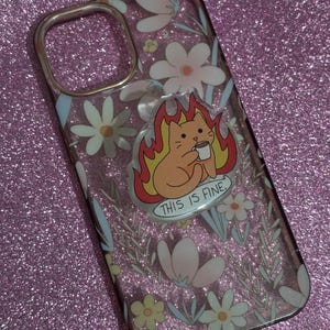 May include: Clear phone case with a floral design and a cartoon cat on fire holding a coffee cup. The text "THIS IS FINE" is below the cat. The case has a silver-colored camera ring.