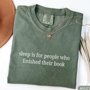 May include: A moss green crew neck t-shirt featuring the white printed text "sleep is for people who finished their book" across the chest. The shirt has a Comfort Colors brand label inside the collar and is paired with a delicate gold necklace.