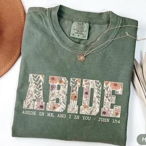 May include: A moss green short-sleeve t-shirt featuring the word ABIDE in large block letters filled with a colorful floral pattern. Below the word, the text reads ABIDE IN ME, AND I IN YOU - JOHN 15:4. The shirt has a crew neckline and a tag.