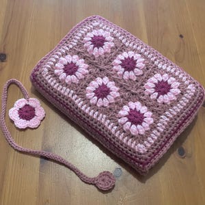 May include: A crocheted book cover and bookmark set. The cover is rectangular, with a floral pattern of pink and burgundy flowers. The bookmark has a matching flower design. The set is made of yarn in shades of pink and burgundy.