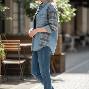 May include: A woman wearing a denim shirt with plaid sleeves and blue jeans. The shirt has a button-down front and two chest pockets. She is also wearing white sneakers. The background shows a street with trees and outdoor seating.