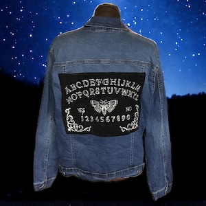 May include: A blue denim jacket with a black patch on the back. The patch features a white embroidered Ouija board with a butterfly and the letters "ABCDEFGHIJKLMNOPQRSTUVWXYZ" and numbers "1234567890".