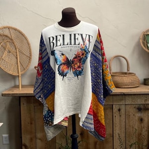 May include: A cream-colored short-sleeve top featuring a large, colorful butterfly graphic with floral patterns on its wings. The text "BELIEVE" is printed in black above the butterfly, with additional text below. The sides feature patchwork fabric.