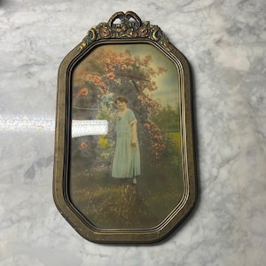 May include: An oval-shaped wall frame with a dark metallic finish and ornate floral detailing at the top. The frame contains a vintage-style print of a person in a light-colored dress standing in a garden with blooming rose bushes and greenery.