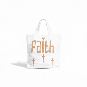 Puede incluir: Un bolso de punto blanco con la palabra Faith bordada en hilo color canela en el centro. Sobre la palabra hay un pequeño corazón color canela y debajo hay tres símbolos de cruz color canela espaciados uniformemente. Tiene dos asas blancas.