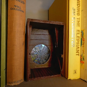 May include: A miniature wooden structure with a round, stained-glass window featuring a spiderweb design. The structure is brown and has a small interior with a wooden floor. It is positioned between two books.