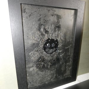 Puede incluir: Una obra de arte enmarcada con una superficie agrietada y un diseño de cara sonriente negra. El marco negro rodea el fondo texturizado y desgastado. La obra de arte tiene una estética oscura y atrevida.