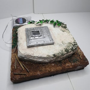 Puede incluir: Un pequeño modelo de búnker de piedra blanco y gris con una escotilla de metal. La escotilla tiene una pequeña ventana cuadrada y está rodeada de remaches de metal. El búnker está colocado sobre una base marrón con vegetación verde y ramitas.