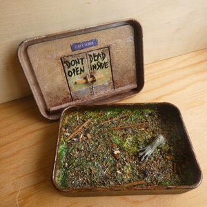 Peut inclure: Diorama miniature dans une boîte rectangulaire. La partie supérieure présente une façade de bâtiment vieillie avec un panneau "CAFETERIA" et une porte avec le texte "DON'T OPEN DEAD INSIDE". La partie inférieure représente une zone herbeuse avec une main.