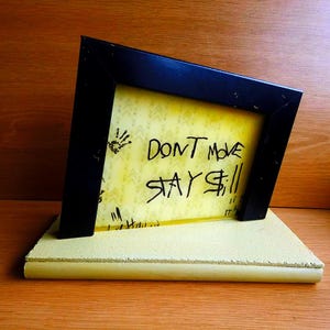 Op de afbeelding: Een zwarte rechthoekige lijst bevat een geel doorschijnend paneel met de handgeschreven zwarte tekst "DONT MOVE STAY STILL" en kleine handafdruk-afbeeldingen. De lijst is gemonteerd op een dikke, lichtgele rechthoekige voet.