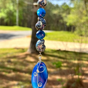 May include: A blue agate pendant with silver wire wrapping, hanging from a silver chain with blue and black patterned beads.