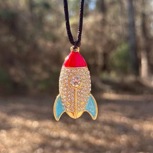 May include: A gold rocket ship pendant necklace. The pendant has a red top, blue fins, and a crystal-encrusted body. It hangs from a black cord. The pendant is approximately 3 cm in length.
