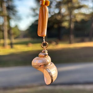 May include: A seashell pendant with a gold-colored metal accent and a string of amber-colored beads. The pendant is a light beige color with a spiral shape. The beads are a mix of shapes and sizes, and the background is blurred.
