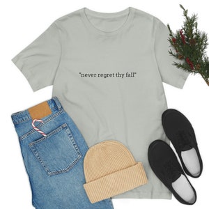May include: A light green t-shirt with the text "never regret thy fall" printed in black on the front. The t-shirt is laid flat on a white background with a pair of blue jeans, a beige beanie, and black sneakers.