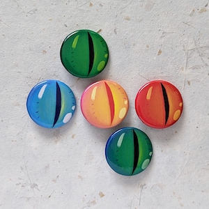 Puede incluir: Cinco botones redondos con ojos de diferentes colores. Los botones son verdes, azules, amarillos, rojos y verdes. Los ojos son todos negros.