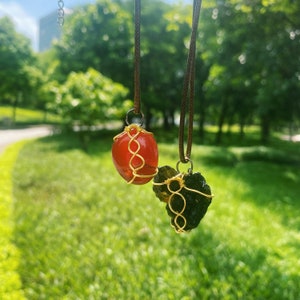 May include: Two gemstone pendants hanging from brown cords. One pendant is a smooth, orange-red stone wrapped in gold-colored wire. The other is a dark green, rough-textured stone, also wire-wrapped. The background is a blurred green park.