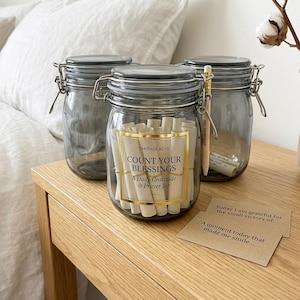 May include: Three gray glass jars with metal latch lids. The center jar features a gold-bordered label reading "SMITHSEALYS COUNT YOUR BLESSINGS A Daily Gratitude & Prayer Jar" and contains rolled paper slips. A pencil and two kraft paper cards are included.