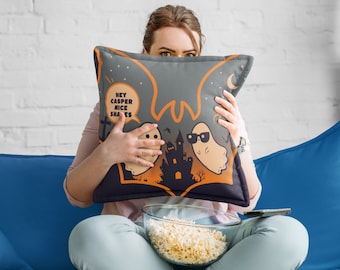 May include: A woman is sitting on a blue couch holding a gray throw pillow with a Halloween-themed design. The pillow features two ghosts, a haunted house, and the text "Hey Casper Nice Shades".